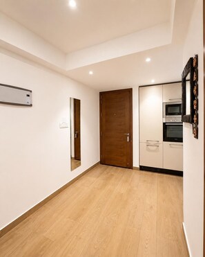 Una habitación con piso de madera, una puerta, aire acondicionado instalado en la pared y una kitchenette con microondas y horno.