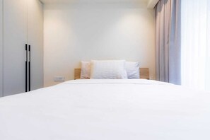 Una habitación de hotel con cama, ropa de cama blanca, cabecera de madera y una ventana con cortinas.