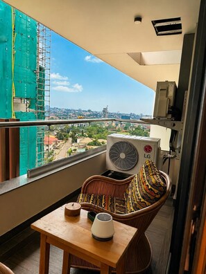 Un balcon donnant sur le paysage urbain, une chaise en osier avec un coussin coloré, une petite table avec une lampe et un appareil de climatisation.