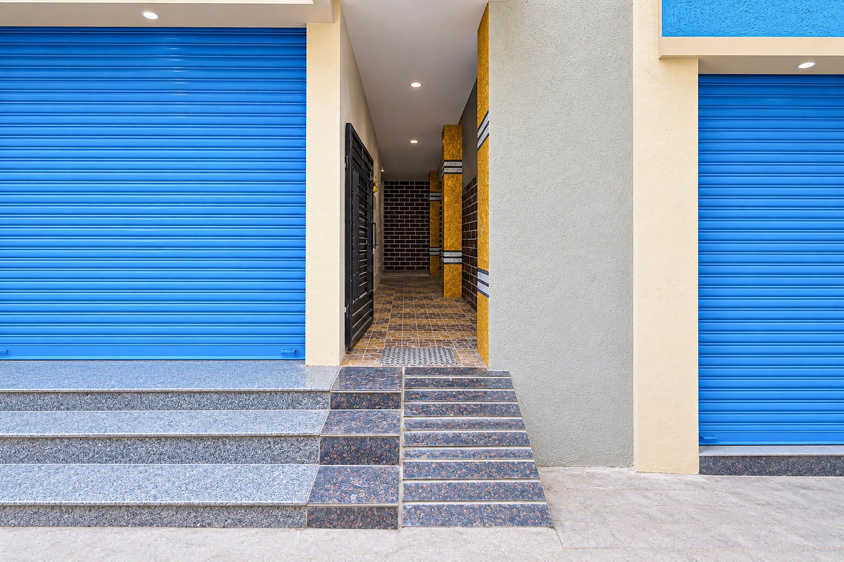 A building entrance with blue roller shutters, a tiled staircase, and a corridor with patterned walls.