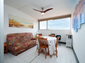 A room with a patterned sofa, a round table with chairs, a ceiling fan, a window with blinds, and a wall-mounted shelf with bottles. - Cabin apartment with a loggia offering sea views (Agde)