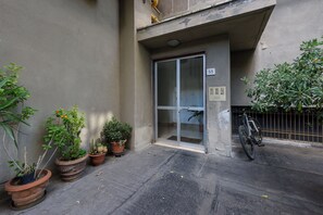 Entrada de un edificio con puerta corredera de vidrio, plantas en macetas y una bicicleta estacionada afuera.