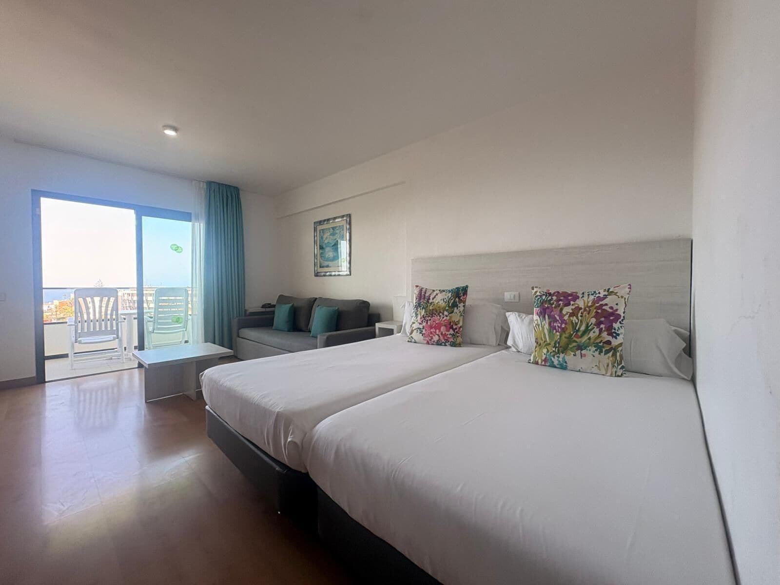 A hotel room with a balcony, a sofa, a bed with floral pillows, and a painting on the wall.