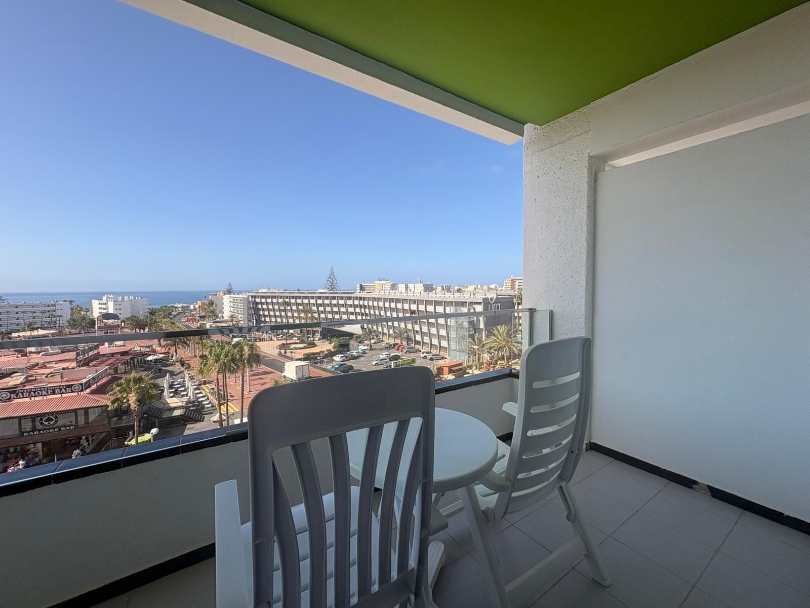 Un balcon avec deux chaises et une table, d’où l’on peut admirer un paysage urbain avec ses bâtiments et un ciel dégagé.
