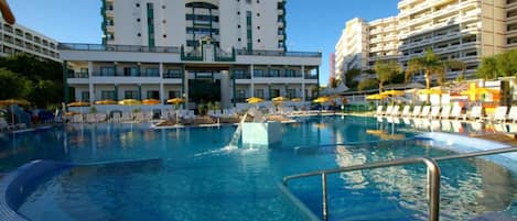 A swimming pool with a water slide, surrounded by lounge chairs and umbrellas, with a hotel building in the background.