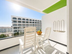 Un balcón con dos sillas blancas, una mesa pequeña con una cesta de frutas y vistas a un edificio de varios pisos.