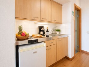 Una cocina compacta con armarios de madera, un refrigerador blanco, una cafetera, una botella de vino y una cesta de frutas.
