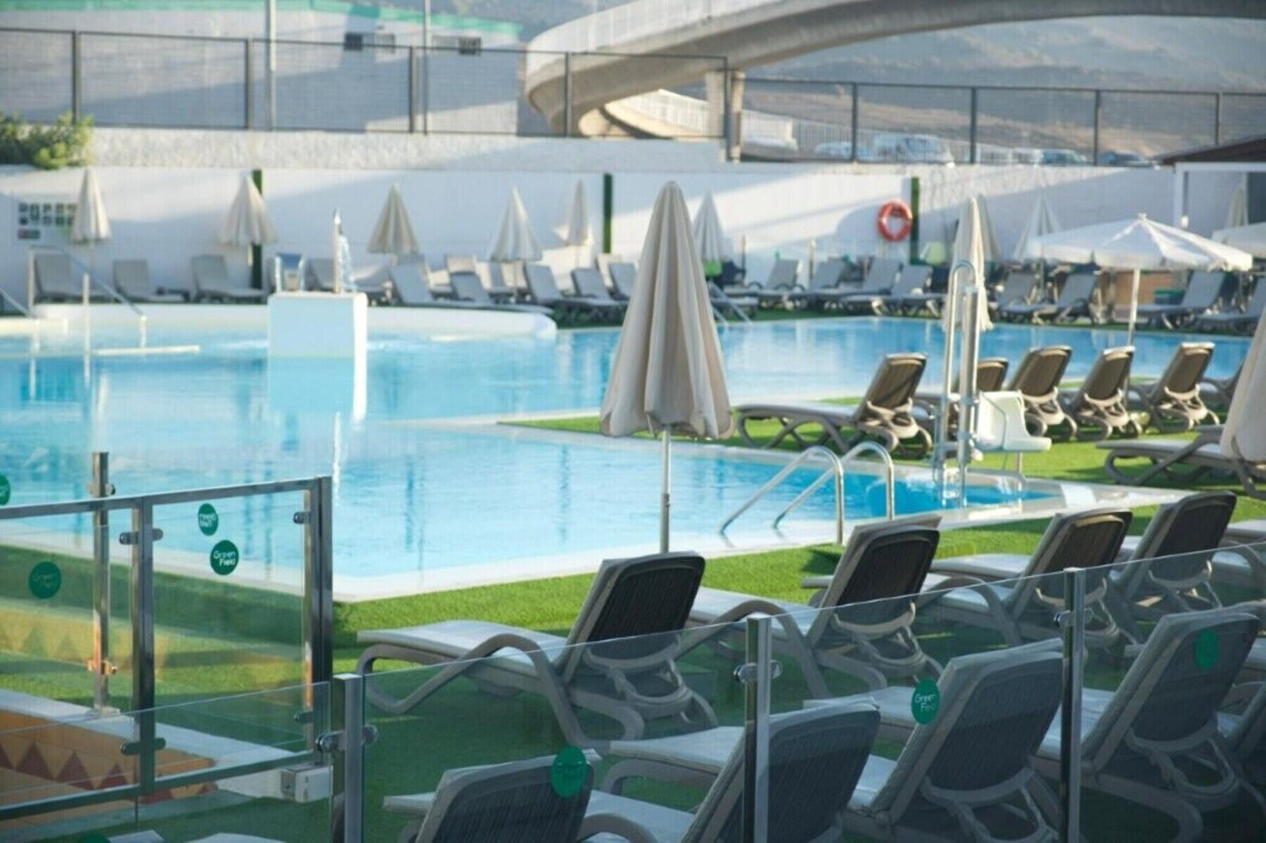 A pool area with lounge chairs and umbrellas, a glass barrier, and a bridge in the background.