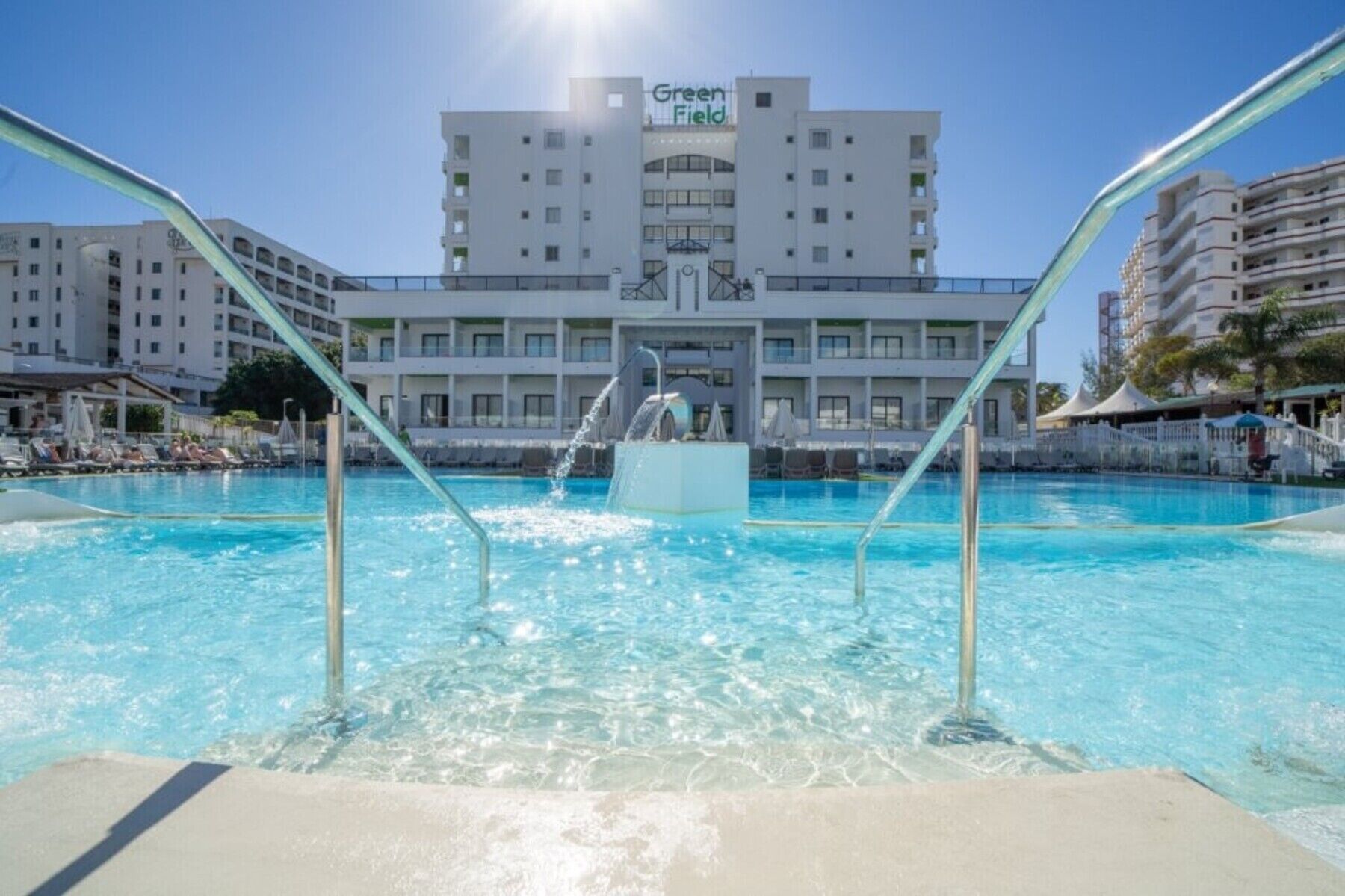 A swimming pool with a water fountain, surrounded by lounge chairs and umbrellas, with a hotel building in the background.