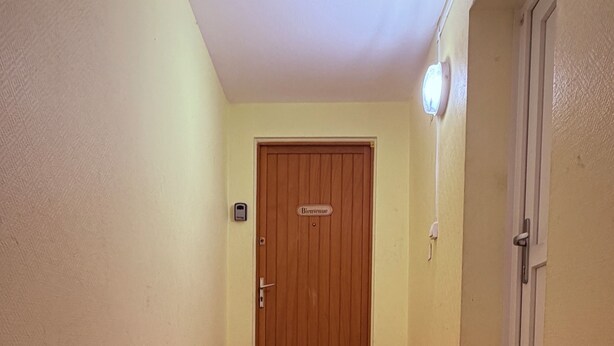 Een gang met een houten deur waarop 'Bienvenue' staat, een lichtschakelaar en een deurbel op de muur.