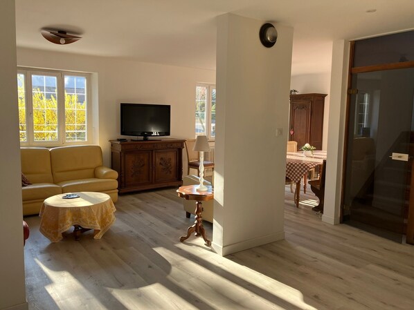 A living room with a sofa, a television, a table with a lamp, and a view of the outside through large windows.