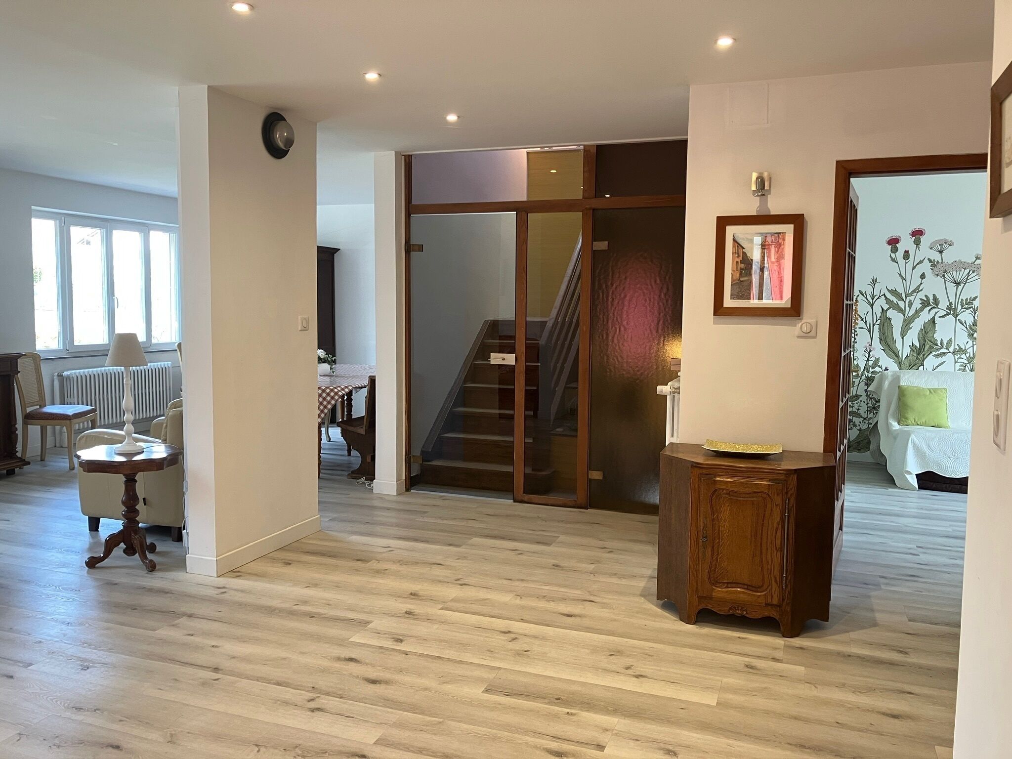 A spacious living area with a wooden floor, a dining table with chairs, a staircase, and a bed with a green pillow.