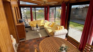A covered outdoor seating area with wooden furniture and a table, connected to a building with wooden beams and a ceiling fan.