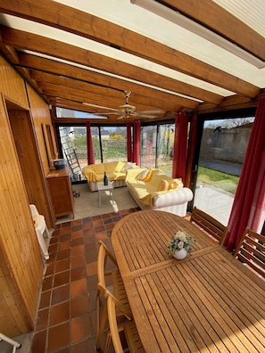A covered outdoor seating area with wooden furniture and a table, connected to a building with wooden beams and a ceiling fan.
