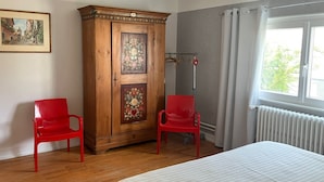 A bedroom with a bed, two red chairs, a wooden wardrobe, a radiator, and a window with curtains.