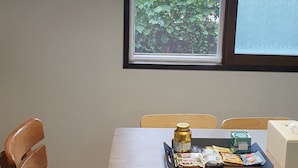 Een eetgedeelte met een tafel, stoelen, een raam met uitzicht op groen en een lijst met een foto aan de muur.