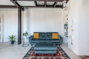 Ein Wohnzimmer mit grauem Sofa, passendem Hocker, einem gemusterten Teppich, einem kleinen Tisch mit Pflanze und einem an der Wand befestigten Telefon.