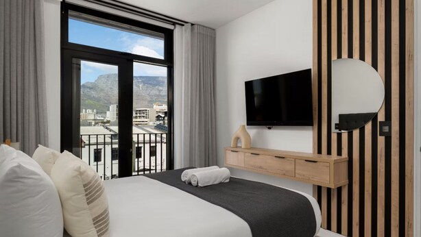 A hotel room with a bed, a TV mounted on the wall, a wooden cabinet, and a view of the city through large windows. - Stylish 1 Bed Secure Spacious Mountain Views (Cape Town)