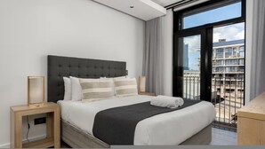 A modern hotel room with a large bed, a bedside table, a nightstand, and a view of the city through a large window. - Stylish 1 Bed Secure Spacious Mountain Views (Cape Town)