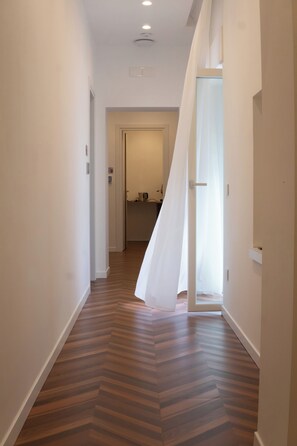 Un pasillo con suelo de madera, una puerta que da a una habitación y una ventana con cortinas de gasa.