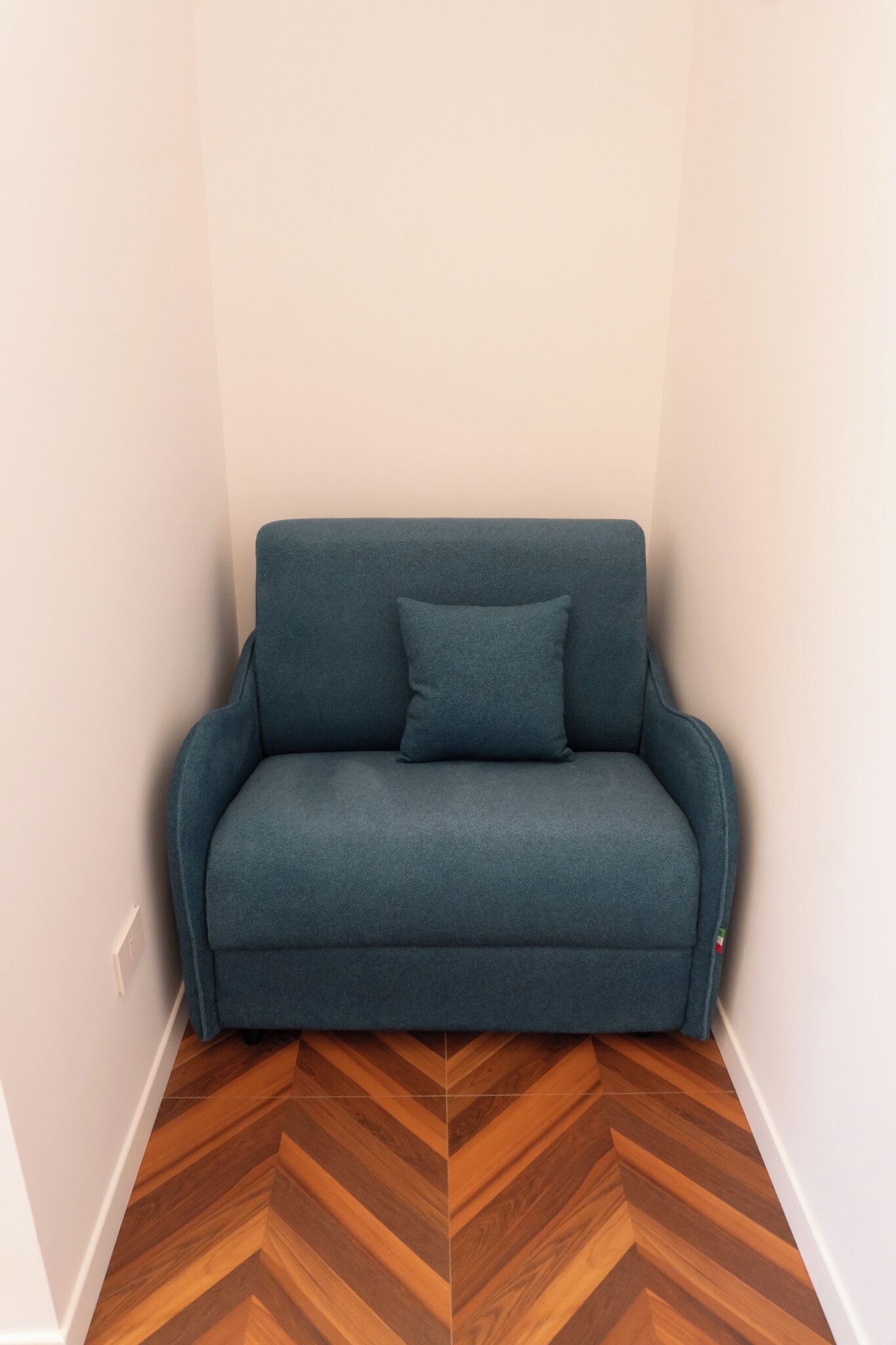 Un piccolo divano scuro con un cuscino in un angolo di una stanza con pavimento in legno e pareti chiare.