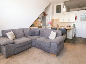 A modern living room with a grey sectional sofa, a kitchen area with white cabinets, and a visible fire extinguisher.
