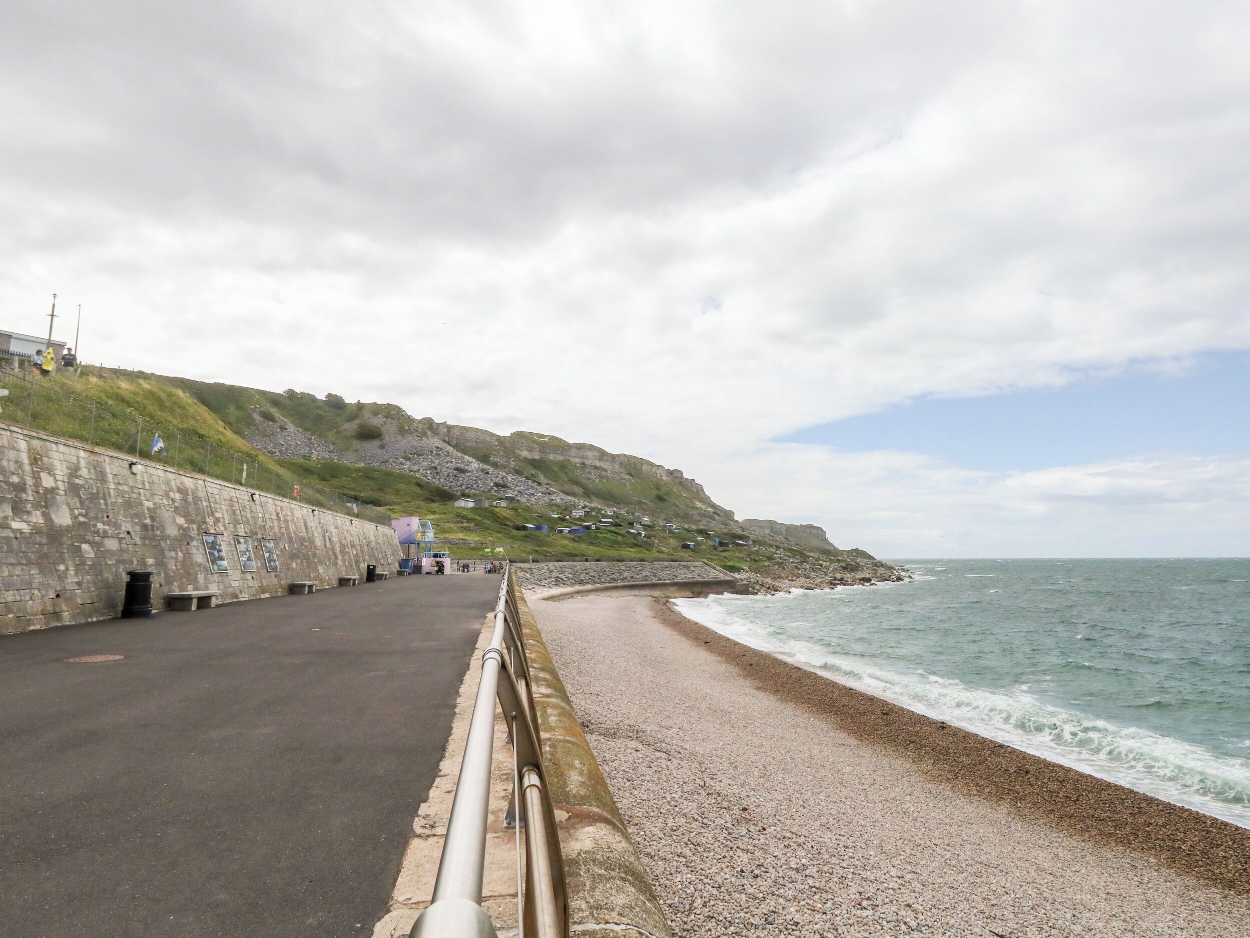 A coastal road with a stone wall, pebbled beach, and ocean waves.