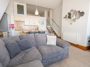 A modern living room with a grey sectional sofa, a dining area with a kitchen in the background, and a staircase leading to an upper floor.