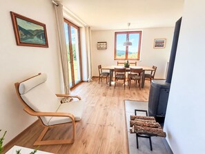 A room with a wooden floor, a dining table with chairs, a sofa, a fireplace, and a large window with curtains.