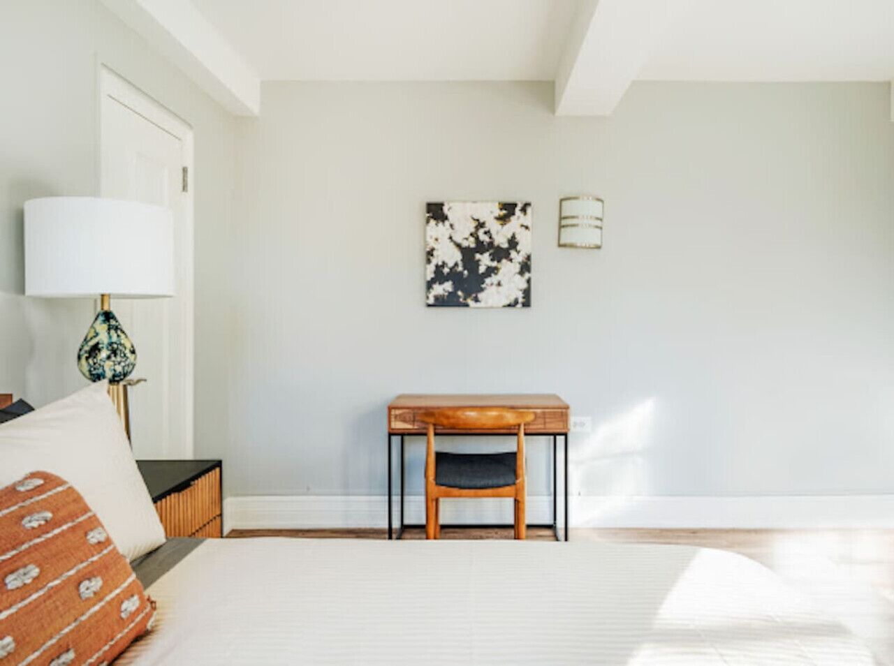 A room with a bed, a table, and a chair. There is a lamp on the table and a picture on the wall.