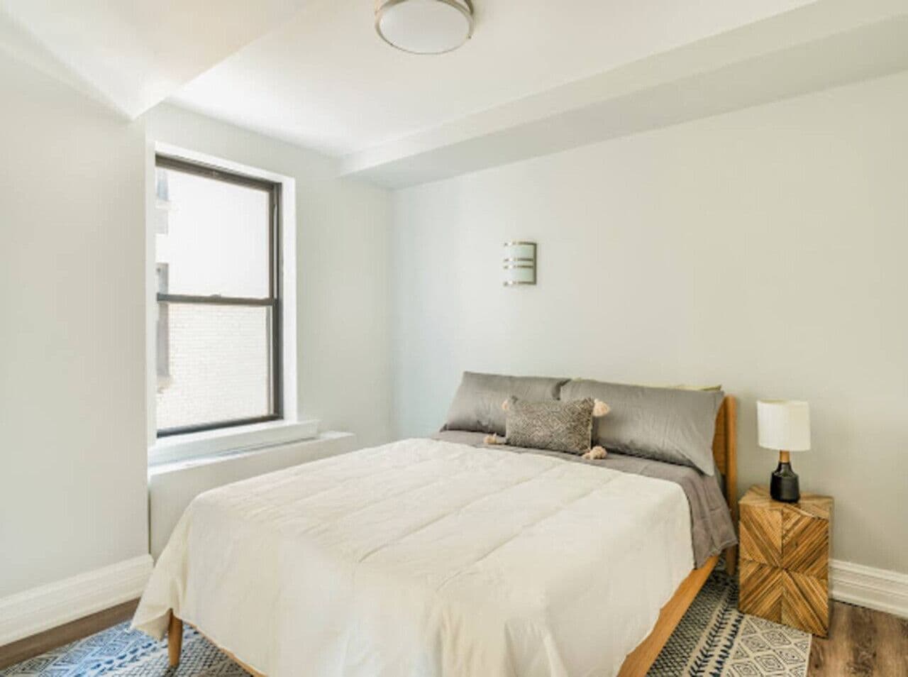 A bedroom with a bed, a window, a nightstand, and a wall-mounted light fixture.