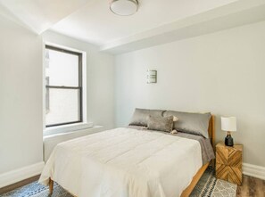Ein Schlafzimmer mit einem Bett, einem Fenster, einem Nachttisch und einer Wandleuchte.