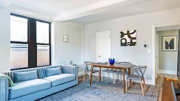 A living room with a sofa, a dining table, and chairs. There is a window with blinds, a door, and a wall-mounted artwork.