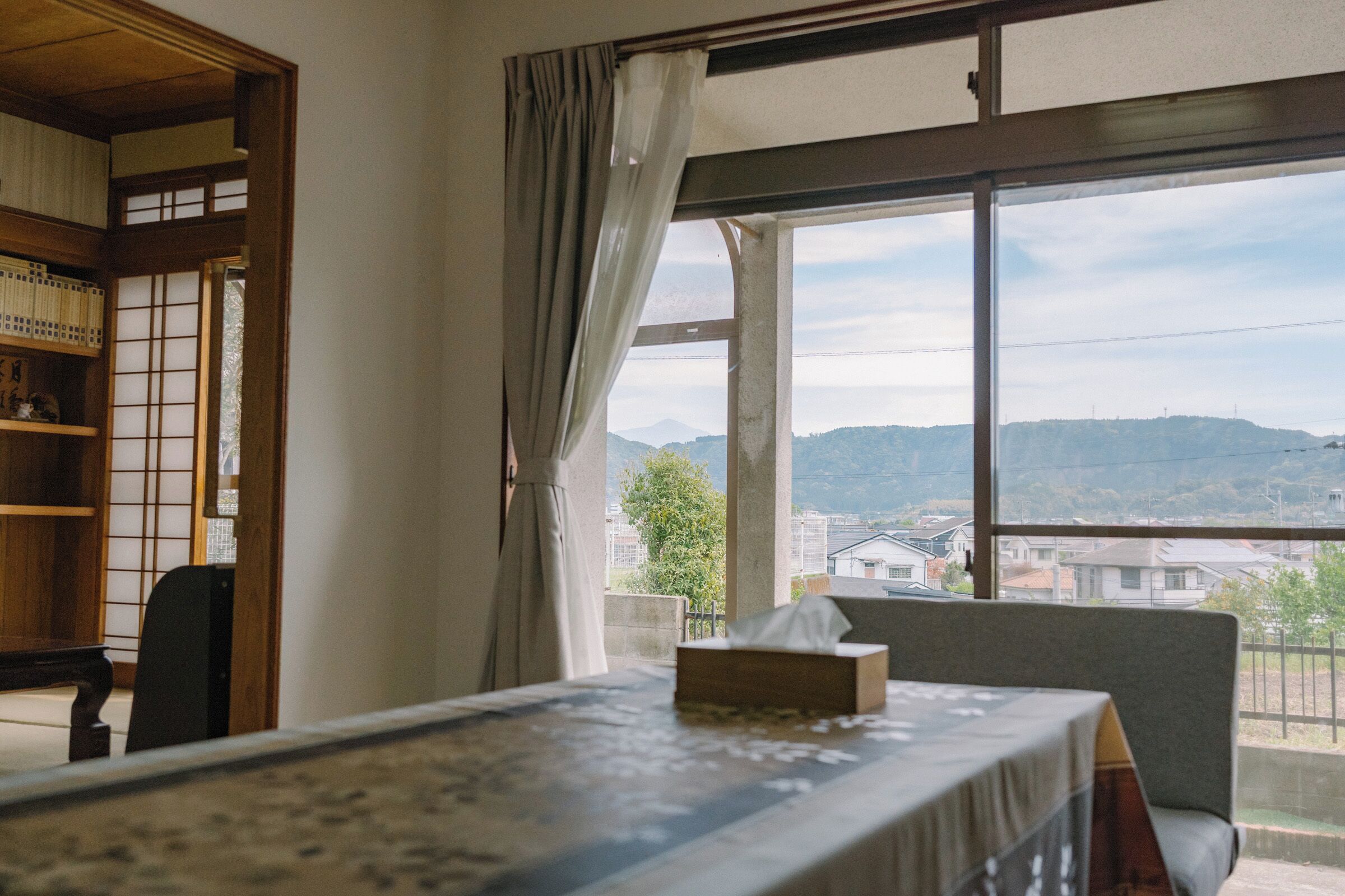 A room with a bed, a chair, a table, and a view of a hilly landscape through a window.