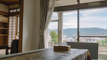 A room with a bed, a chair, a table, and a view of a hilly landscape through a window.