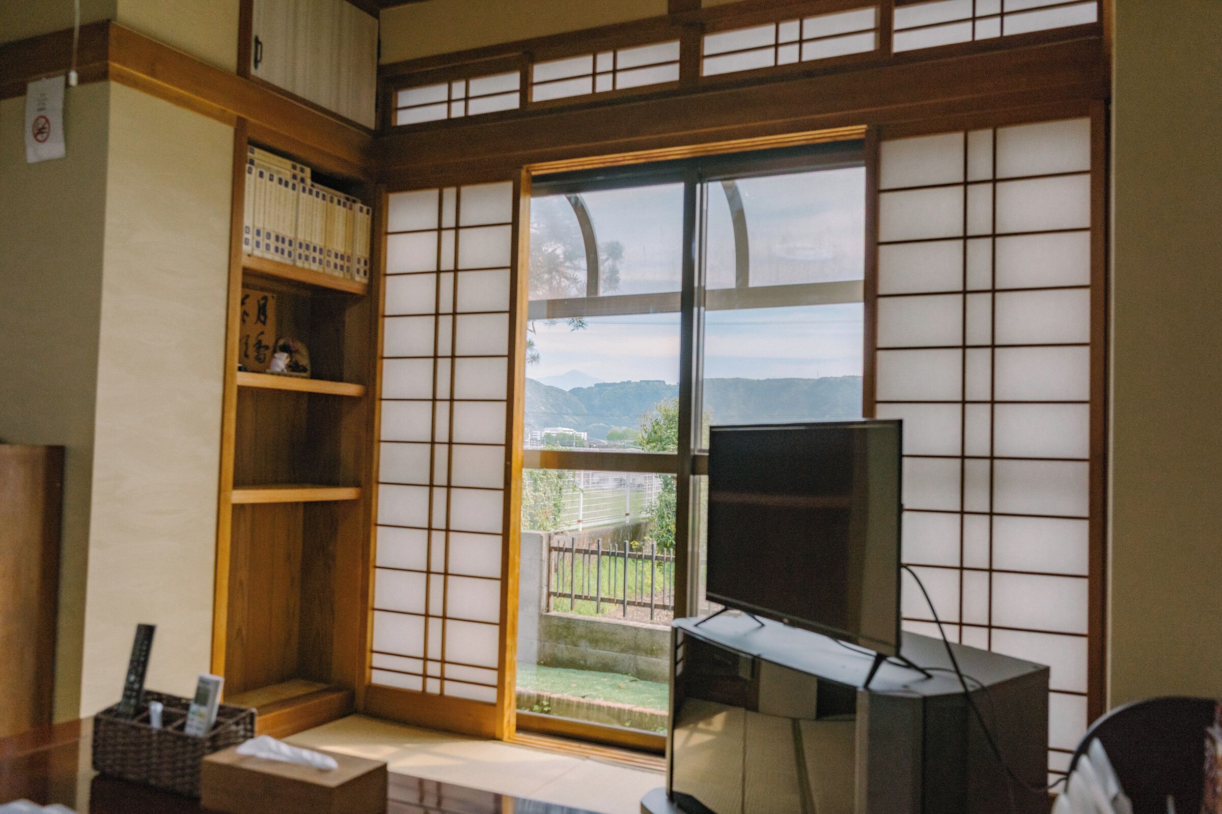 Ein traditioneller japanischer Raum mit Tatami-Bodenbelag, einer Schiebetür mit Aussicht, einem Fernseher und einem Bücherregal.