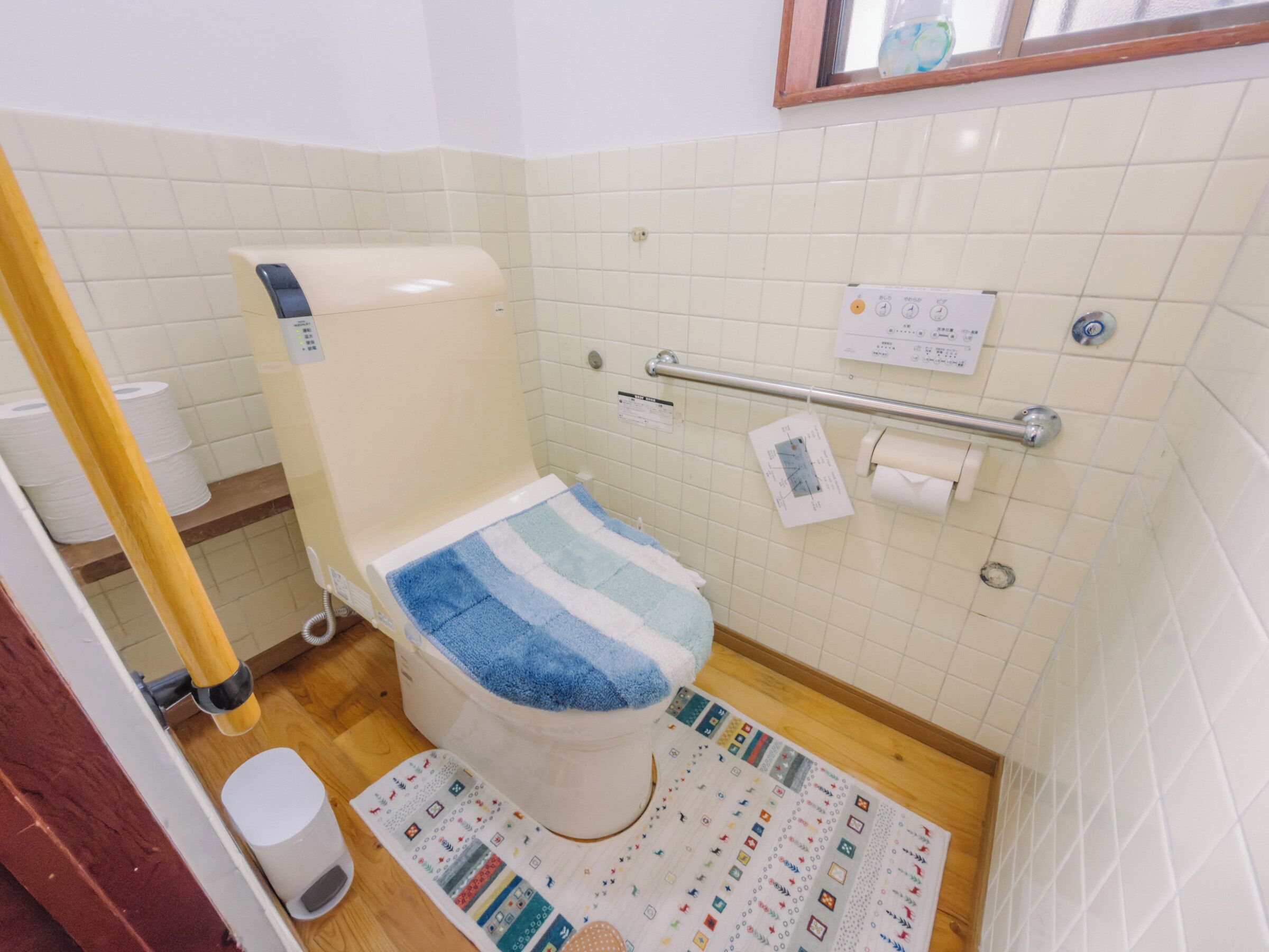 A bathroom with a toilet, a handrail, a roll of toilet paper, and a patterned mat on the floor.