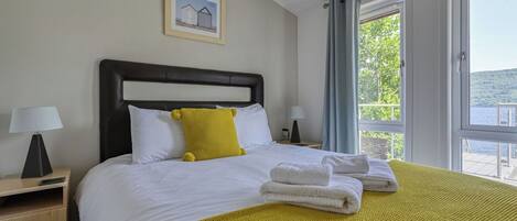 Habitación de hotel con cama, mesita de noche, lámpara, un cuadro en la pared y ventana con vistas a zonas verdes.