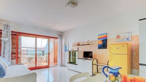 Uma sala ampla com uma porta de vidro deslizante que dá para uma varanda, uma mesa de jantar com toalha de mesa colorida e uma cozinha com geladeira amarela.