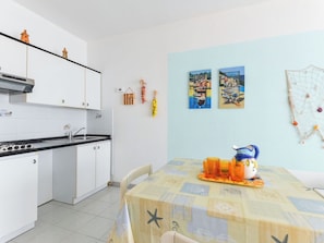 Eine Küche mit weißen Schränken, einer schwarzen Arbeitsplatte und einer gefliesten Spritzwand. Ein Esstisch mit gelber Tischdecke und orangefarbenen Gläsern ist ebenfalls vorhanden.