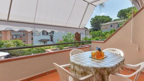 Terraço com mesa posta para dois, uma cadeira branca e uma pequena mesa com jarra e copos.