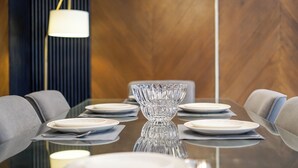 A modern dining table with a reflective surface, surrounded by grey upholstered chairs, set with white plates and a clear glass bowl centerpiece.