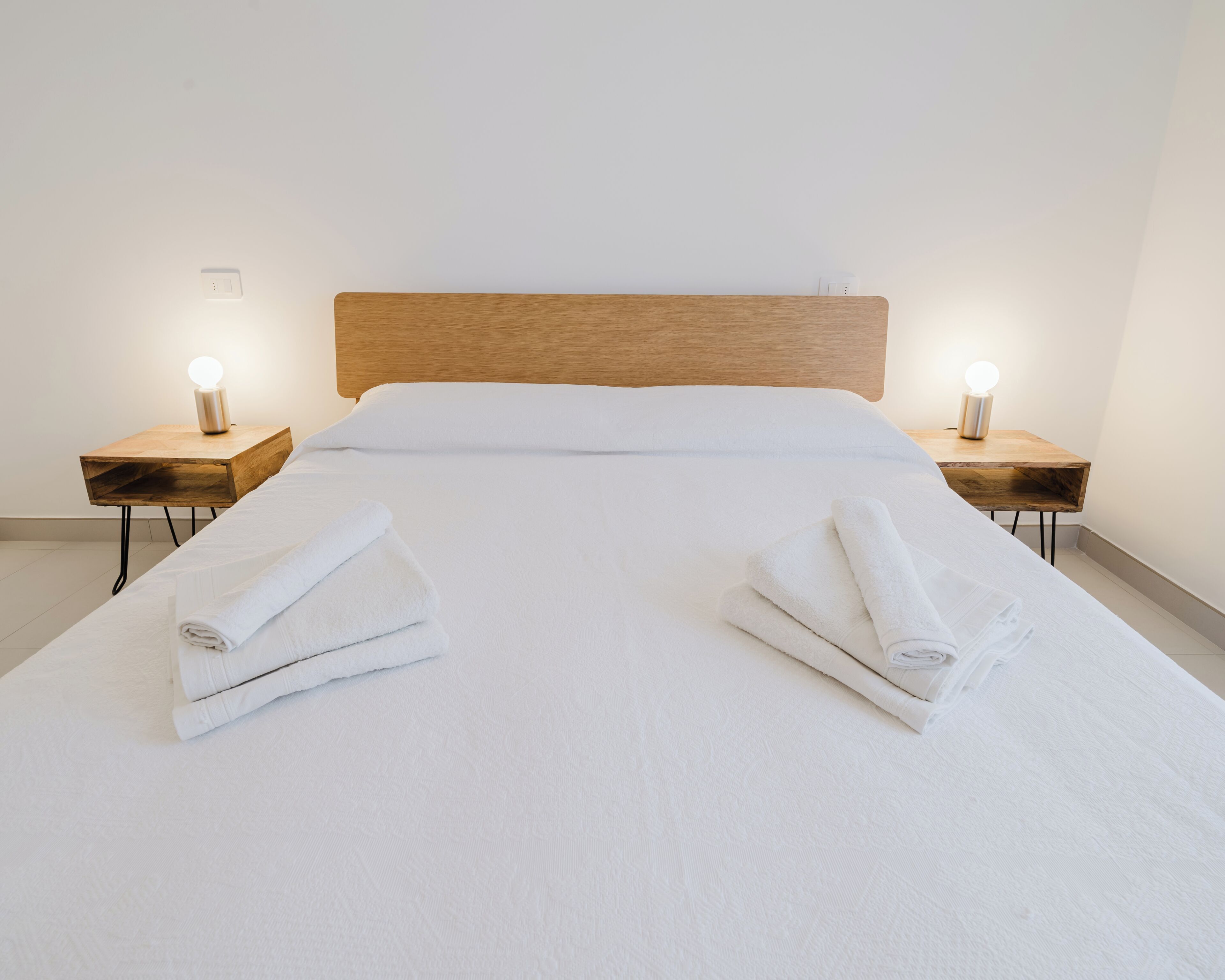 A neatly made bed with white linens, two bedside tables with lamps, and folded towels on the bed.