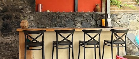 Un espace bar extérieur doté d’un mur en pierre, d’un plafond en bois et d’un comptoir en pierre. On y trouve quatre tabourets de bar noirs et une seule chaise verte avec un coussin.
