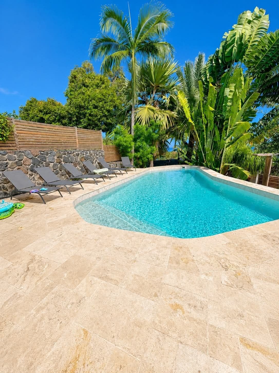 A rectangular swimming pool with clear blue water, surrounded by a stone deck and a wooden fence.