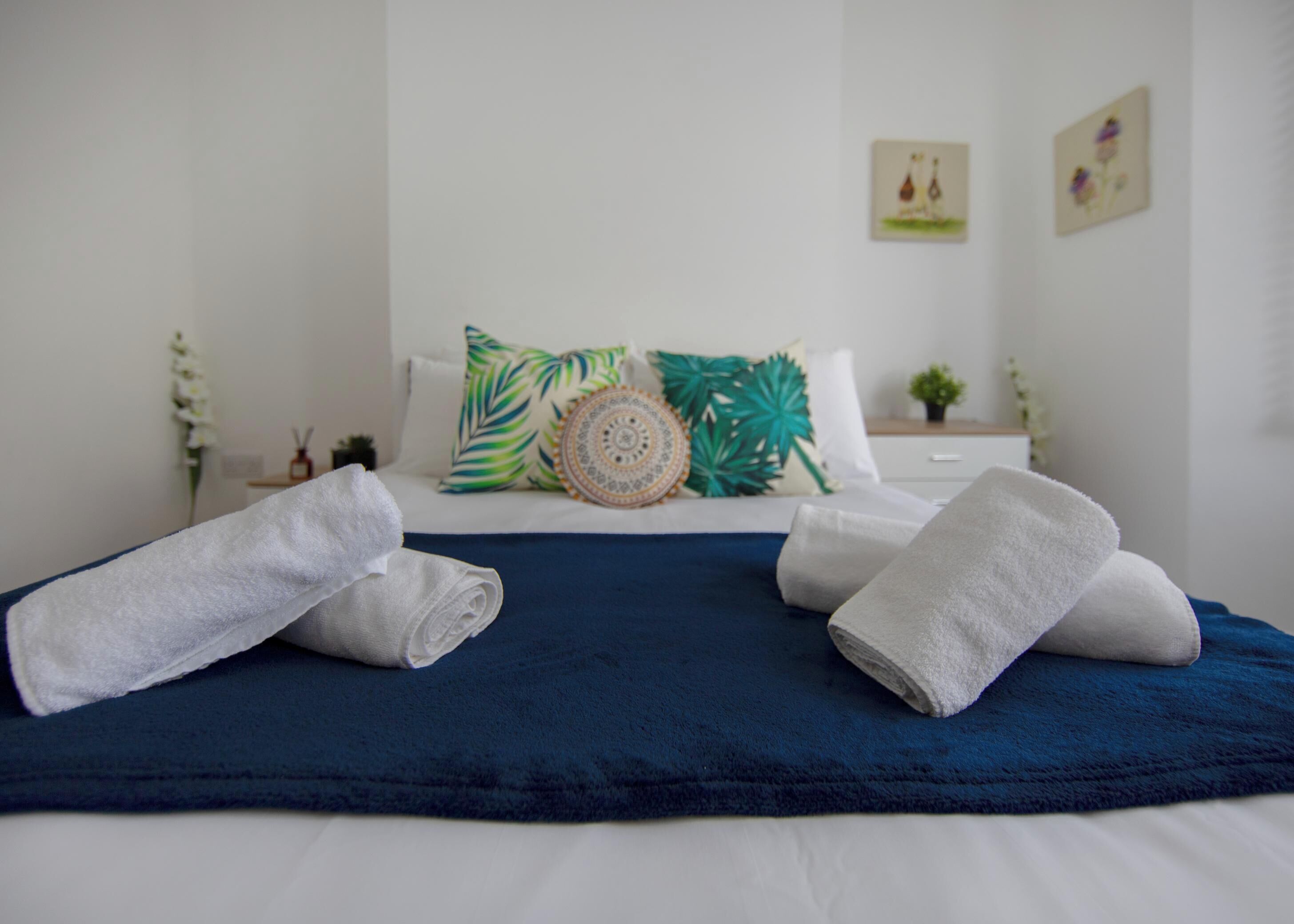 A neatly made bed with a blue bedspread and two rolled towels, a nightstand with a small plant, and a wall with two framed pictures.
