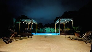 Un espace piscine extérieur doté de deux coins salon couverts, chacun agrémenté de guirlandes lumineuses et de chaises.