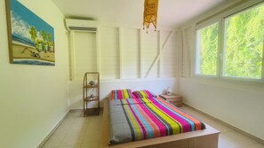 A bedroom with a bed, a nightstand, a lamp, a window with a view of greenery, and a painting on the wall. - Villa Les Alpinias: A Serene Oasis with a Private Pool (Petit-Bourg)