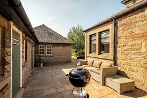 A stone building with a green door labeled 'THE WOODSHED,' a patio with a grill, and a wicker sofa.