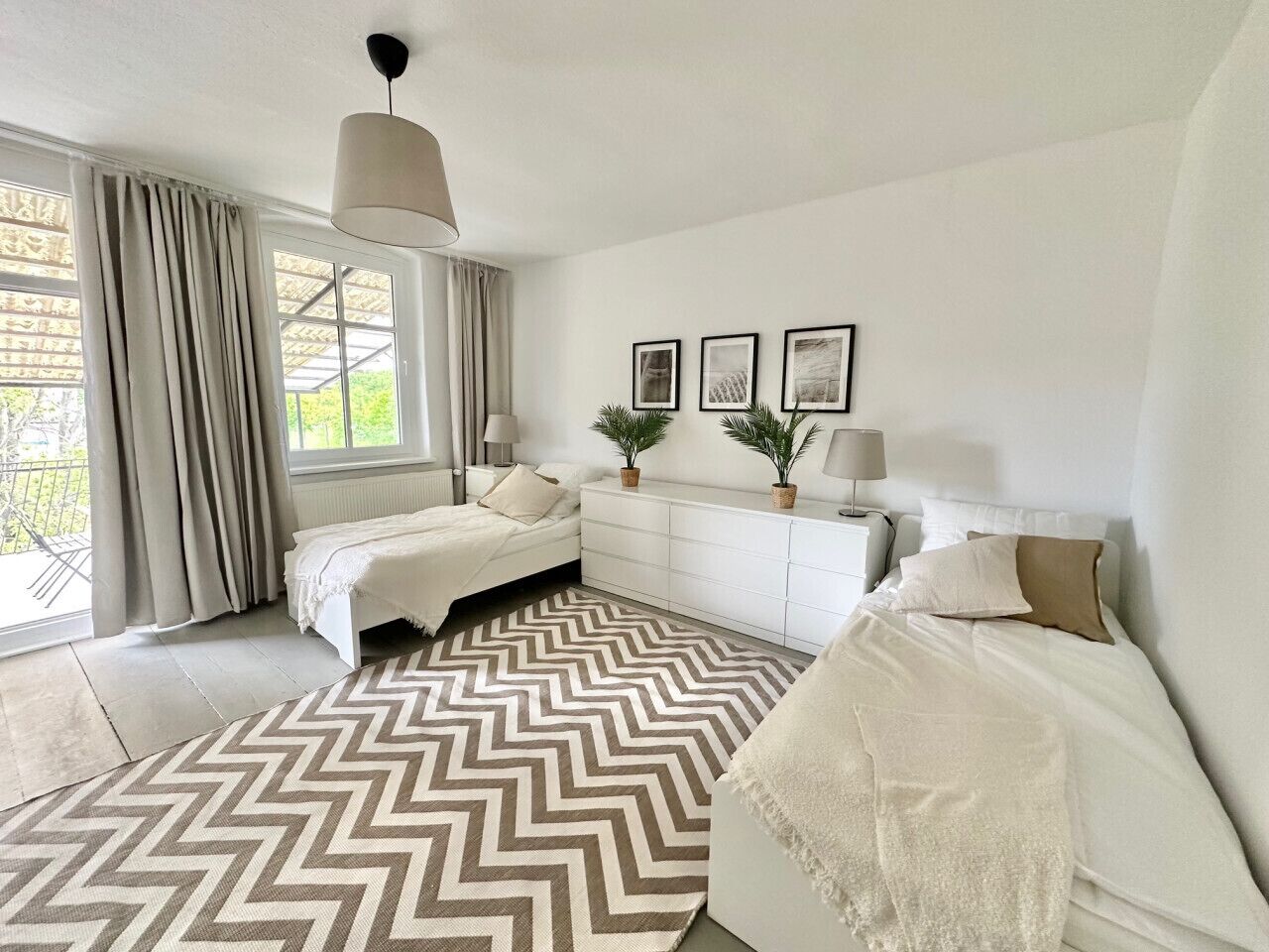 A bedroom with two beds, a chevron-patterned rug, a white dresser, and a large window with curtains.